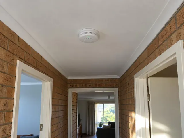 Ceiling-mounted smoke alarm in a brick hallway with white trim in Lake Munmorah